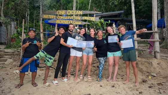 A group of volunteers with Monica & staff members taking a picture at our sign board by the beach